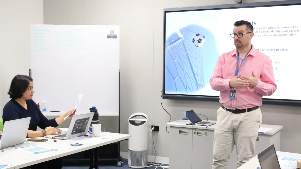 Robert Williams, wearing glasses and in a pink shirt stands in front of a presentation screen. He is talking to a workshop attendee seated at a table, with papers and a laptop in front of her